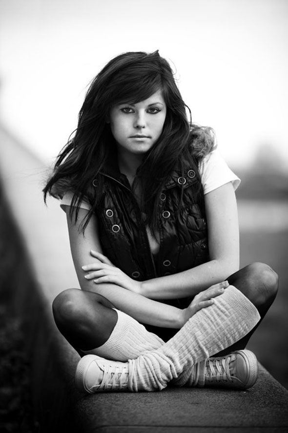 Portret młodej kobiety w czarno-białej tonacji, siedzącej ze skrzyżowanymi nogami, ubranej w białą koszulkę, czarną kamizelkę i getry, z poważnym wyrazem twarzy.