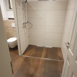 Usługi remontowo-budowlane - Nowoczesna łazienka z brązowymi płytkami podłogowymi, kabiną prysznicową typu walk-in z deszczownicą i toaletą w tle, widok zza otwartych drzwi.