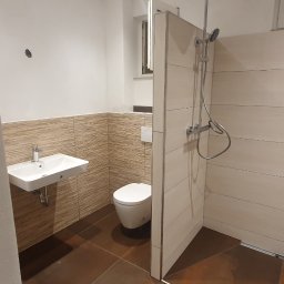 Usługi remontowo-budowlane - Nowoczesna łazienka po remoncie z brązowymi płytkami podłogowymi, umywalką na ścianie, toaletą i kabiną prysznicową z deszczownicą.