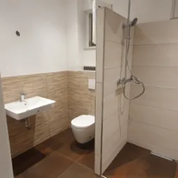 Nowoczesna łazienka po remoncie z brązowymi płytkami podłogowymi, umywalką na ścianie, toaletą i kabiną prysznicową z deszczownicą.