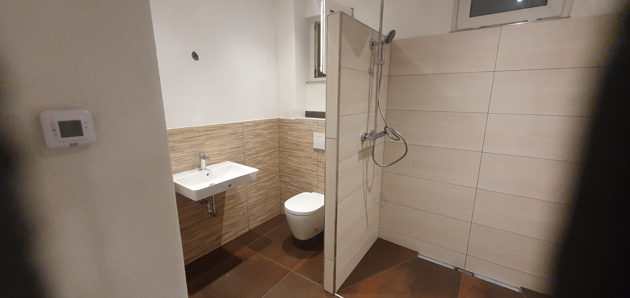 Nowoczesna łazienka po remoncie z brązowymi płytkami podłogowymi, umywalką na ścianie, toaletą i kabiną prysznicową z deszczownicą.