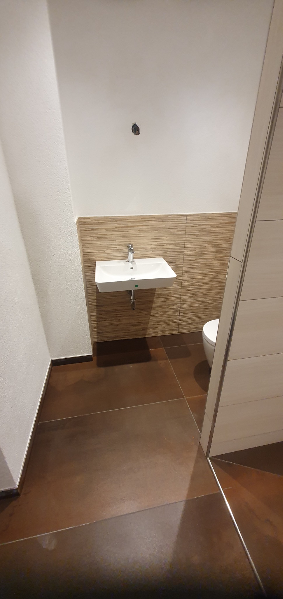 Nowoczesna łazienka z brązową podłogą, wiszącą białą umywalką i beżową mozaiką na ścianie, widoczny fragment białej toalety i drzwi z jasnego drewna.