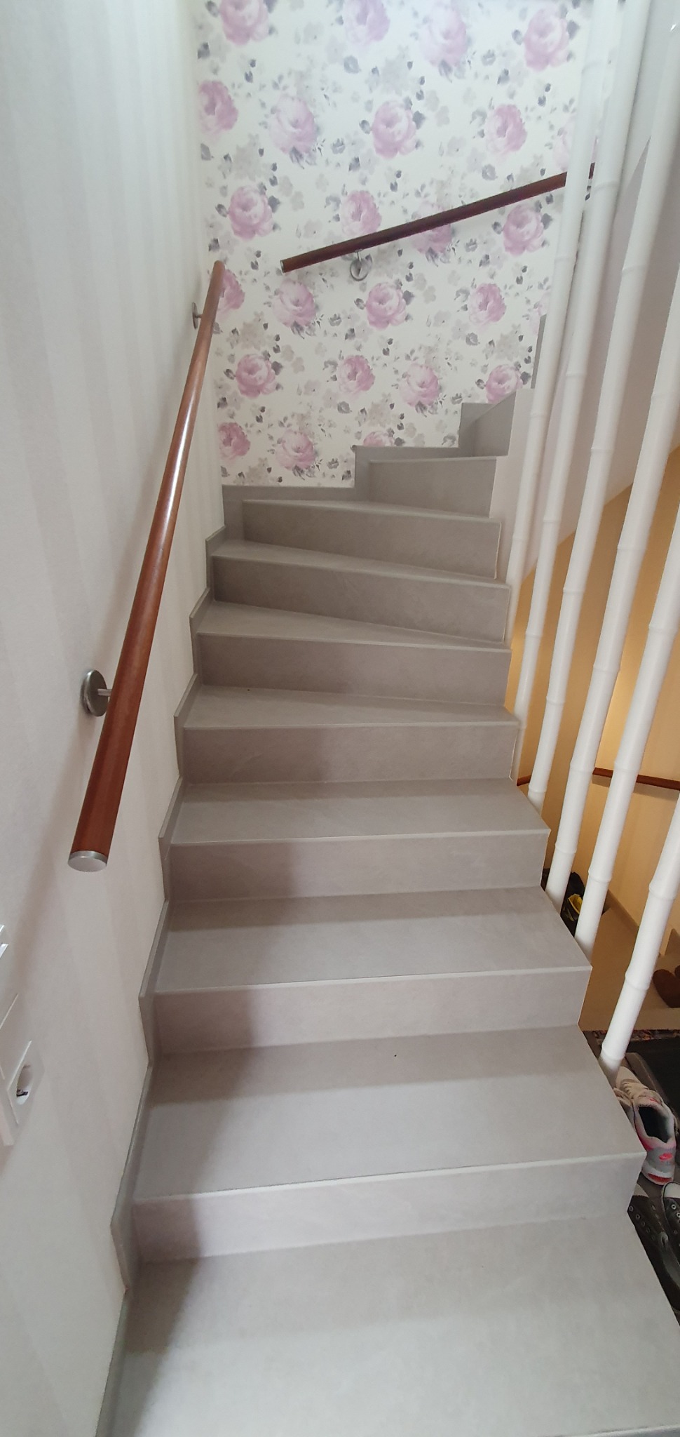 Schody wewnętrzne wykończone w jasnych odcieniach szarości, z drewnianą poręczą po lewej stronie i ozdobnymi, białymi słupkami po prawej, na tle ściany w kwiatowy wzór.