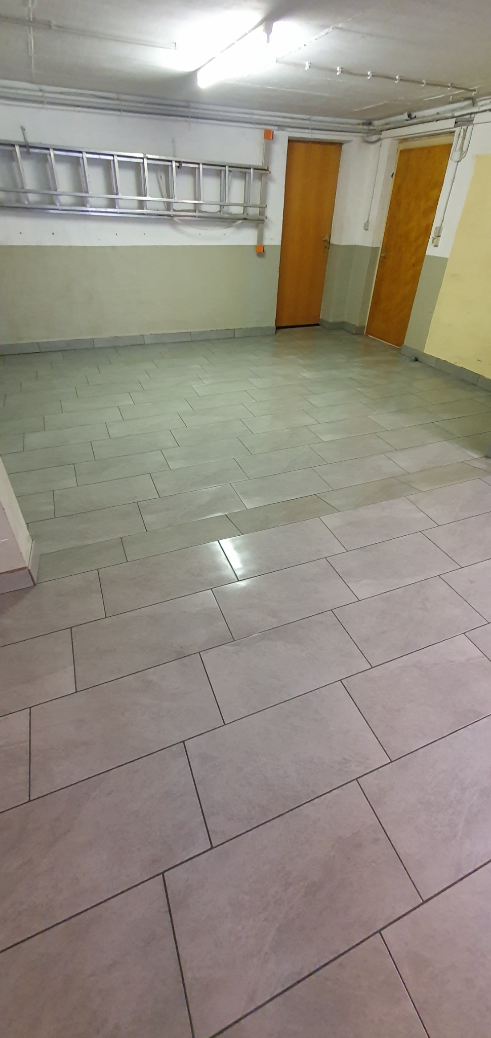 Ułożone, prostokątne, szare płytki podłogowe w pomieszczeniu z dwoma drewnianymi drzwiami i aluminiową drabiną wiszącą na ścianie.