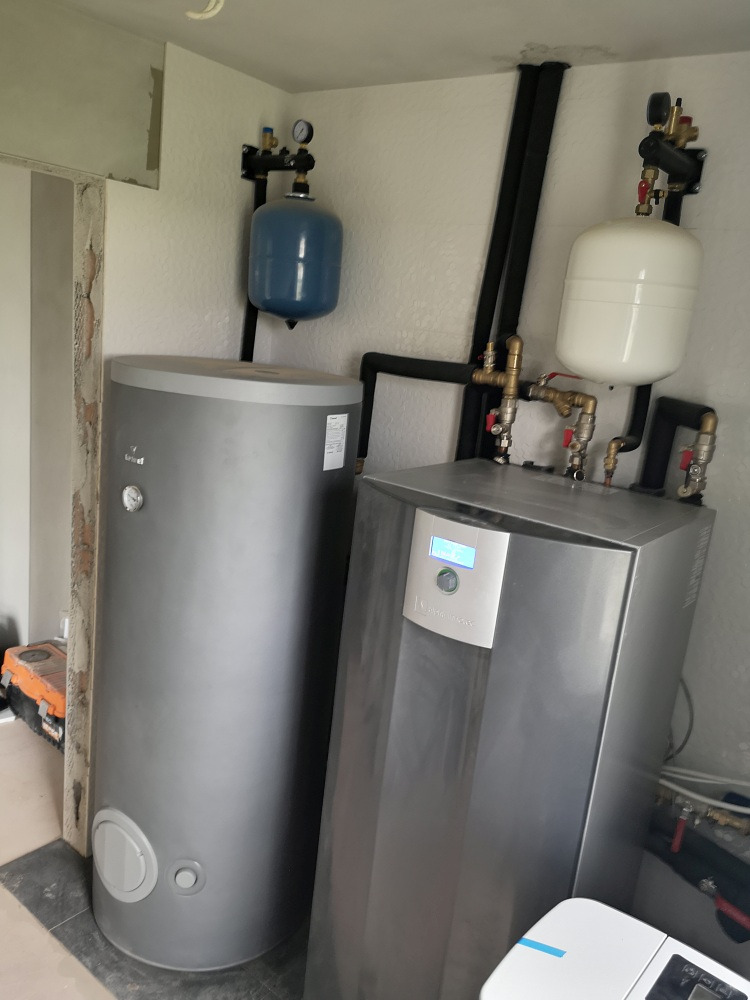 Zainstalowana pompa ciepła Alphainnotec obok pionowego zasobnika CWU o dużej pojemności 290 litrów, z widocznymi elementami instalacji hydraulicznymi i czarnymi rurami, w pomieszczeniu w trakcie...