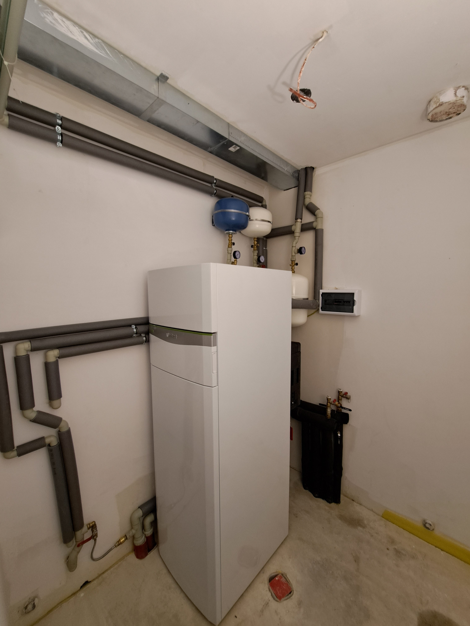 Biała gruntowa pompa ciepła Vaillant z wbudowanym zasobnikiem cwu, widoczne rury w izolacji termicznej, elementy instalacji hydraulicznej i elektrycznej w surowym pomieszczeniu.