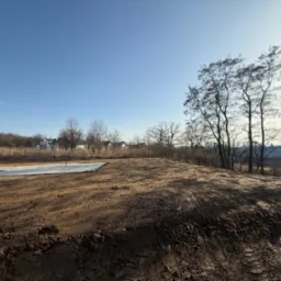 Przygotowany teren pod budowę z widoczną wylewką fundamentową w tle, otoczony drzewami i zabudową jednorodzinną w oddali, błękitne niebo.