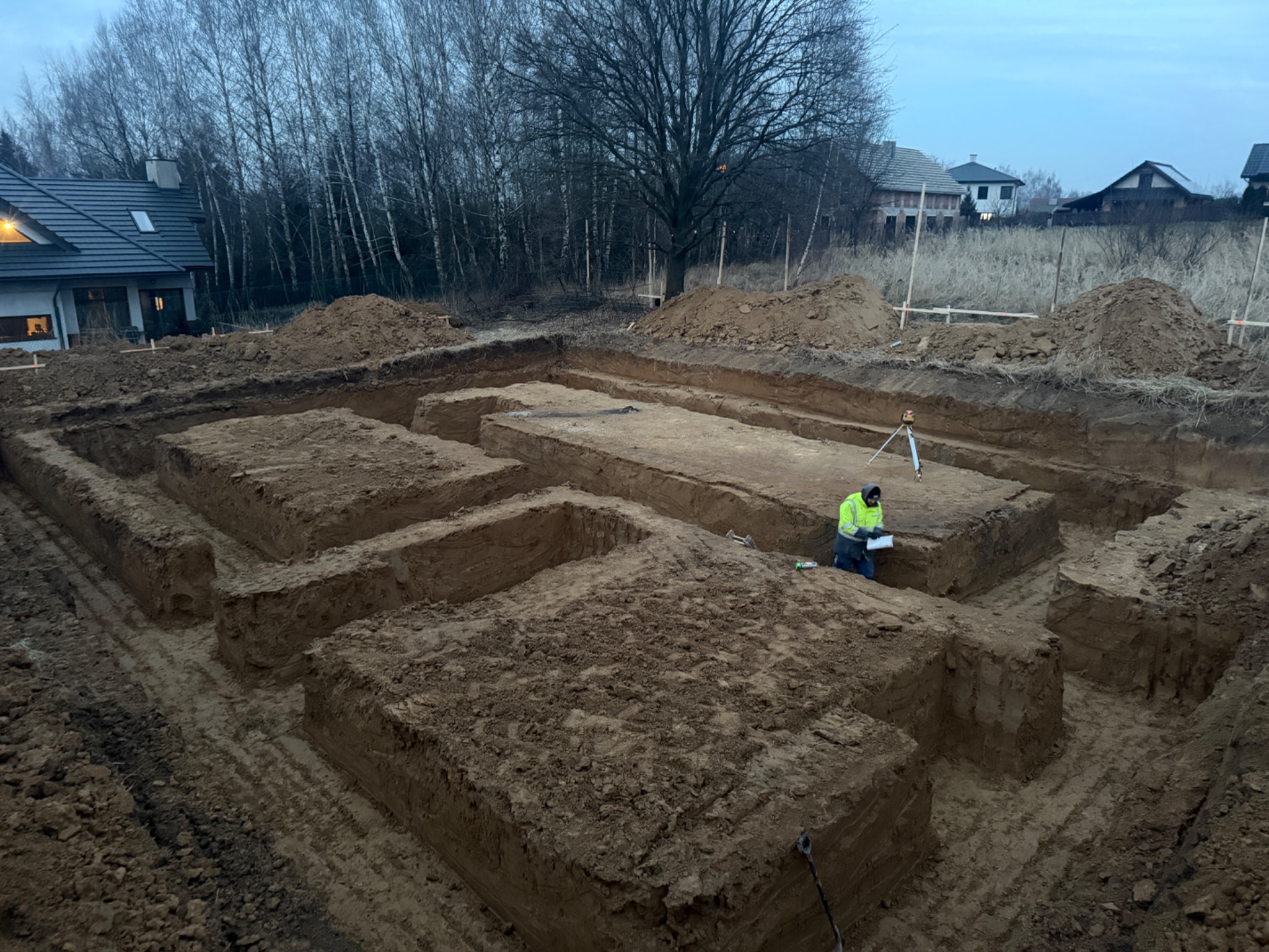 Szeroki kadr na wykopane fundamenty pod budynek, z widocznymi rowami, pryzmami ziemi i pracownikiem w jaskrawej kamizelce mierzącym teren za pomocą niwelatora laserowego. W tle domy i nagie drzewa.