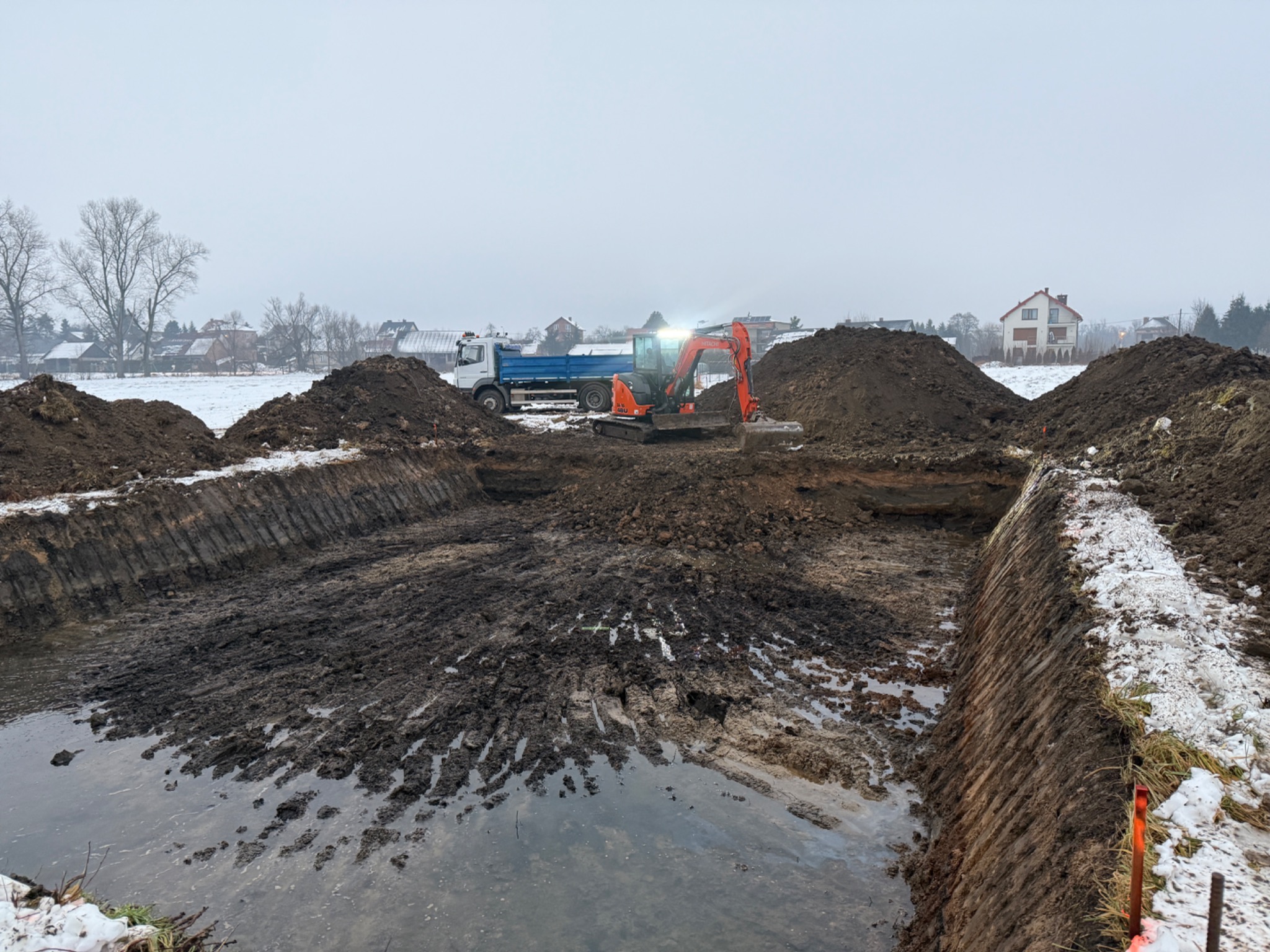 Pomarańczowa koparka Hitachi, oświetlona reflektorem, pracuje w wykopie wypełnionym wodą, z hałdami ziemi po bokach i ciężarówką w tle, w pochmurny dzień.