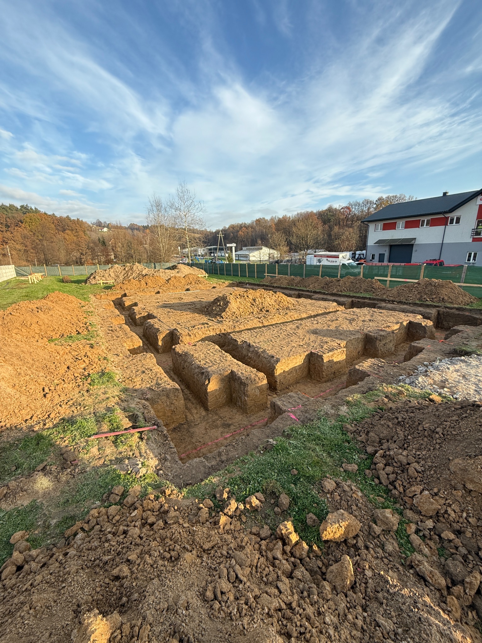 Przygotowany teren pod budowę z wykopanymi rowami pod fundamenty, widoczne wytyczone linie, w tle budynek i drzewa pod błękitnym niebem.
