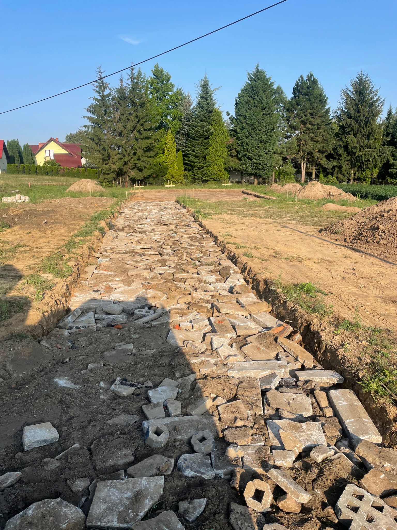 Przygotowanie podłoża drogi z gruzu i ziemi w wykopie, widoczne drzewa i dom w tle.