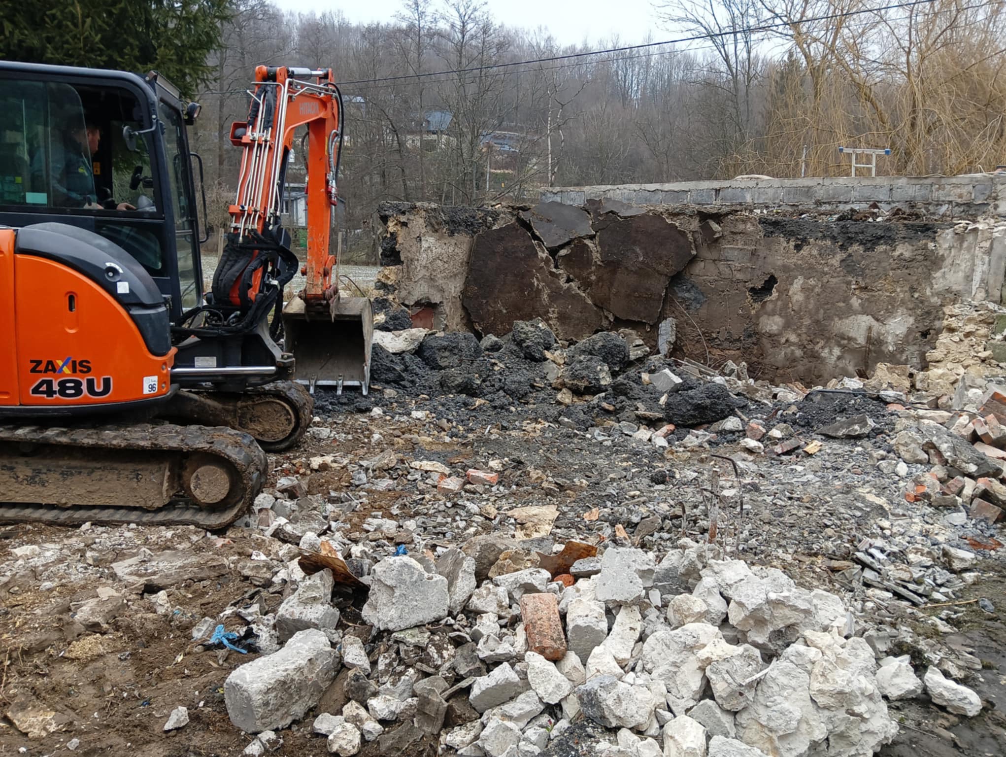 Pomarańczowa minikoparka Zaxis 48U podczas prac rozbiórkowych muru z cegieł i betonu, z gruzem na pierwszym planie i drzewami w tle.