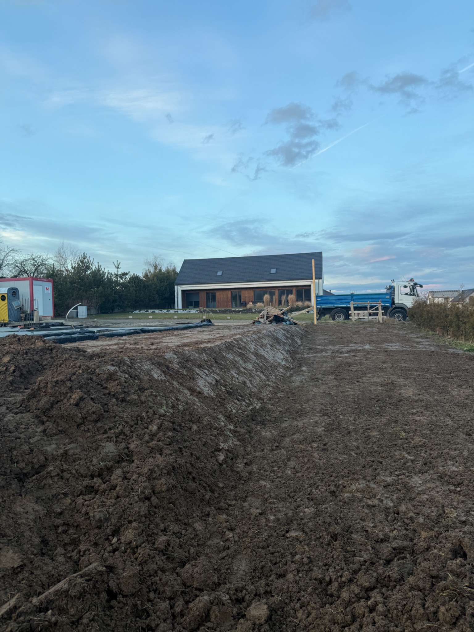 Przygotowany teren pod budowę domu jednorodzinnego, widoczny wykop, czarna folia drenażowa i ciężarówka z niebieską skrzynią ładunkową w tle.