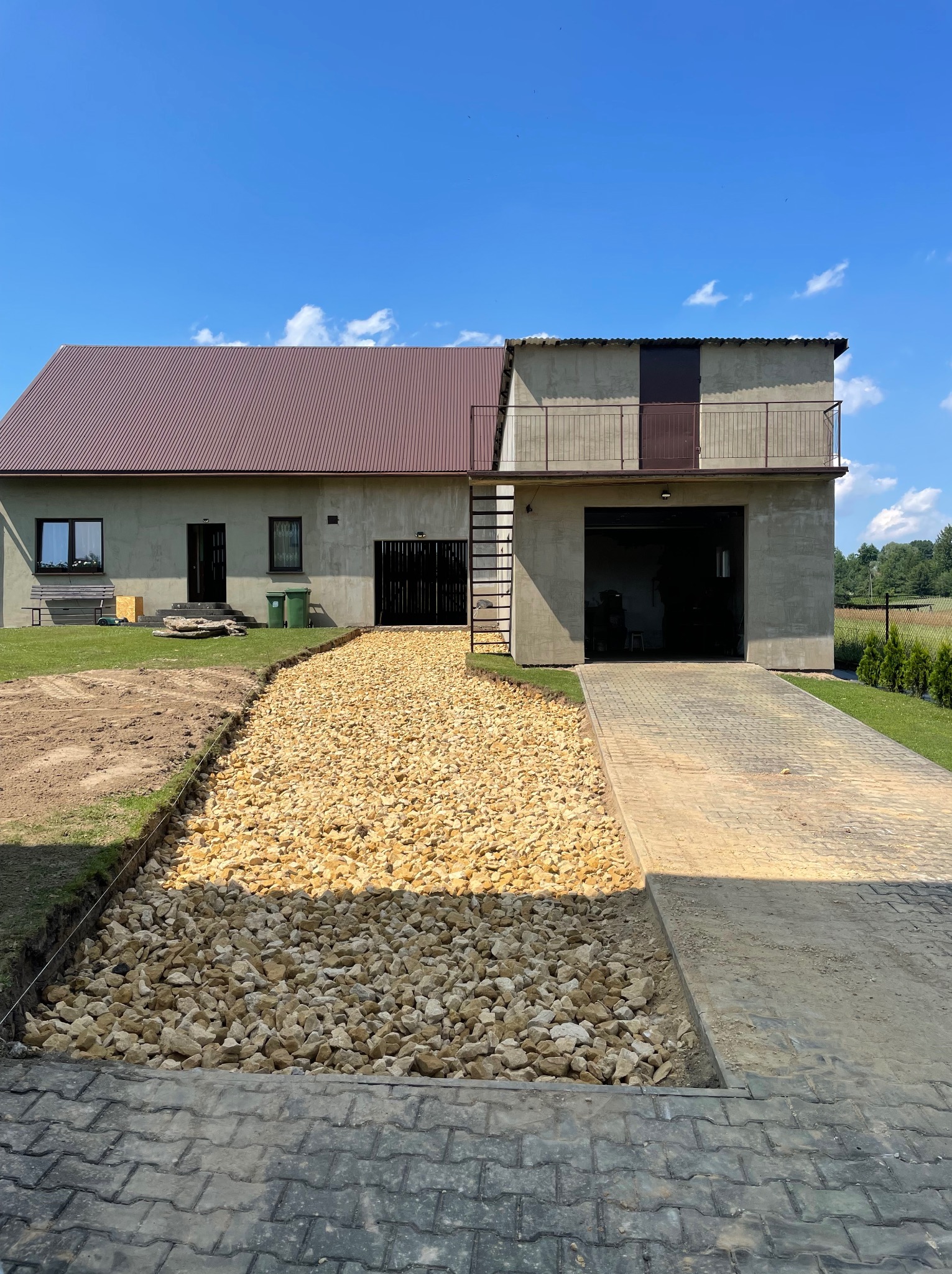 Kruszywo kamienne wysypane na przygotowanym podłożu przed domem jednorodzinnym z garażem, widok na fragment trawnika i kostkę brukową.