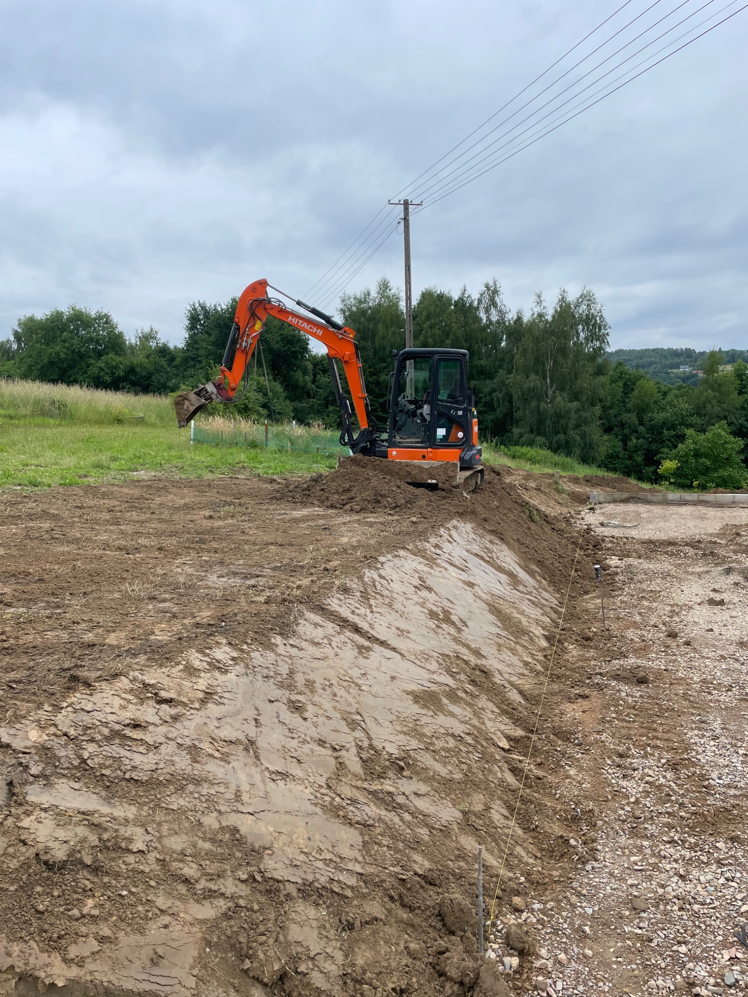 Pomarańczowa mini koparka Hitachi pracuje na placu budowy, wykonując wykop w ziemi. Widoczne napięte sznurki wyznaczające linię wykopu oraz ukształtowany teren z drzewami w tle.