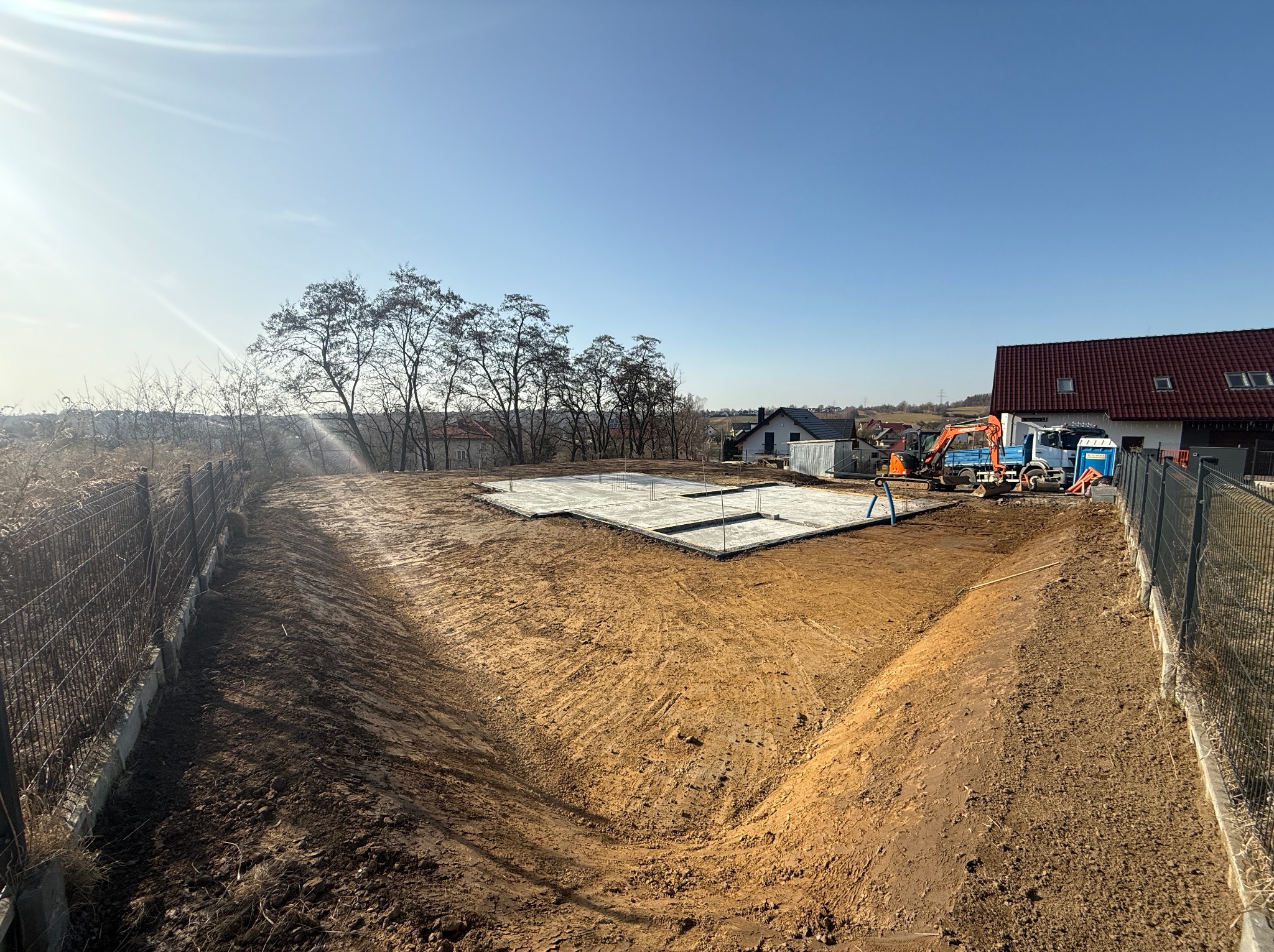 Przygotowany teren pod budowę z wykopem i wylanymi fundamentami, widoczny mały pomarańczowy koparka i biała ciężarówka zaparkowane obok przenośnej toalety, w tle domy jednorodzinne i błękitne niebo.