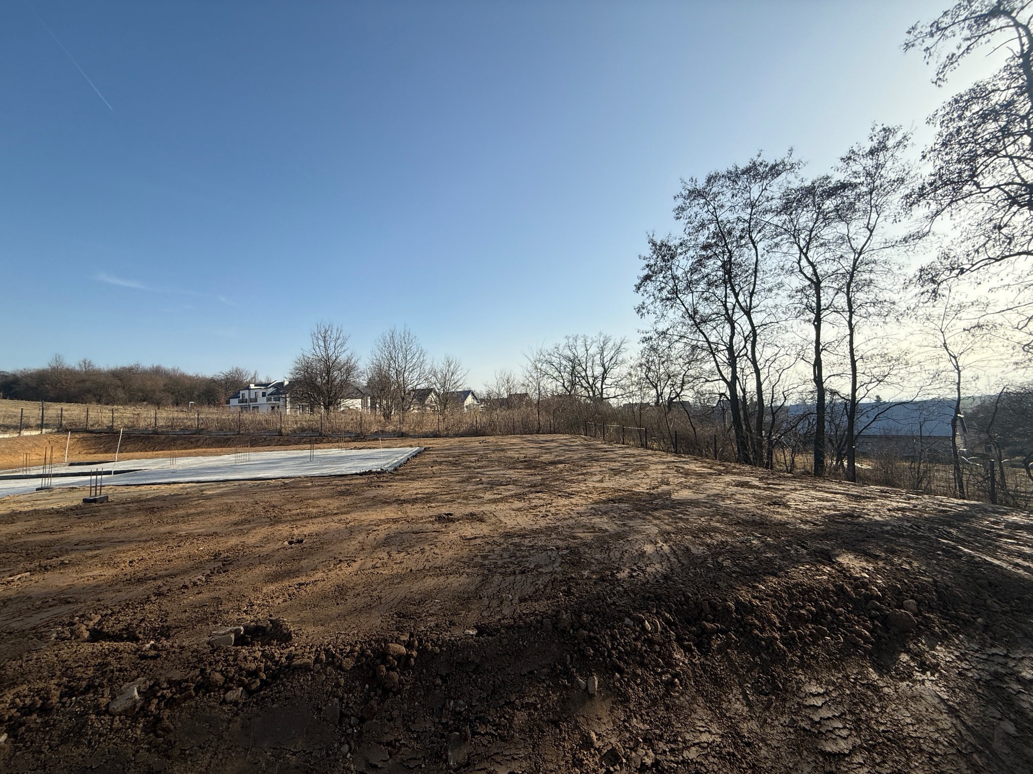 Przygotowany teren pod budowę z widoczną wylewką fundamentową w tle, otoczony drzewami i zabudową jednorodzinną w oddali, błękitne niebo.