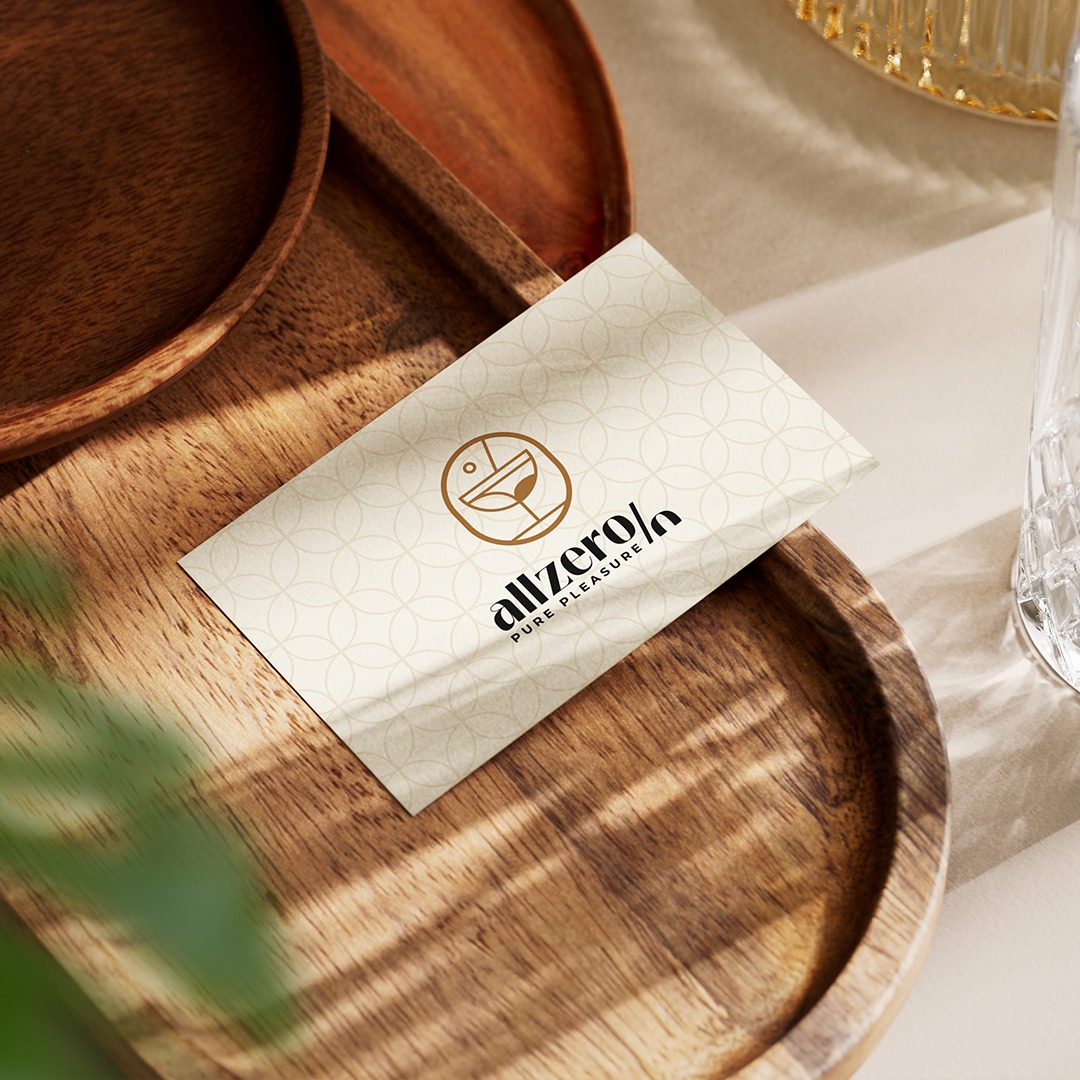 Wizytówka z logo i nazwą 'allzero% PURE PLEASURE' na drewnianej tacy, obok szklanki z kryształowym wzorem, w ciepłym oświetleniu.