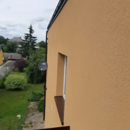 Fragment elewacji domu w odcieniach beżu z widocznym oknem, brązowym parapetem i anteną satelitarną na tle zieleni ogrodu i zabudowań.