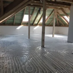 Montaż ogrzewania podłogowego w poddaszu: białe rurki ułożone na folii z nadrukiem producenta, widoczne drewniane krokwie i słupy konstrukcyjne.