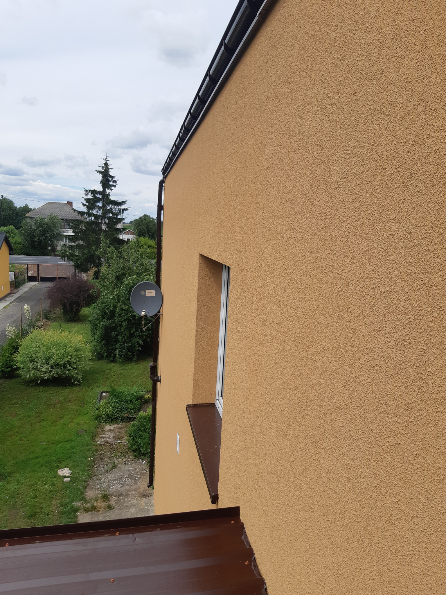 Fragment elewacji domu w odcieniach beżu z widocznym oknem, brązowym parapetem i anteną satelitarną na tle zieleni ogrodu i zabudowań.