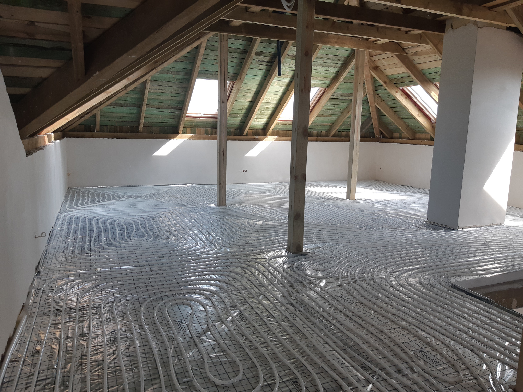Montaż ogrzewania podłogowego w poddaszu: białe rurki ułożone na folii z nadrukiem producenta, widoczne drewniane krokwie i słupy konstrukcyjne.