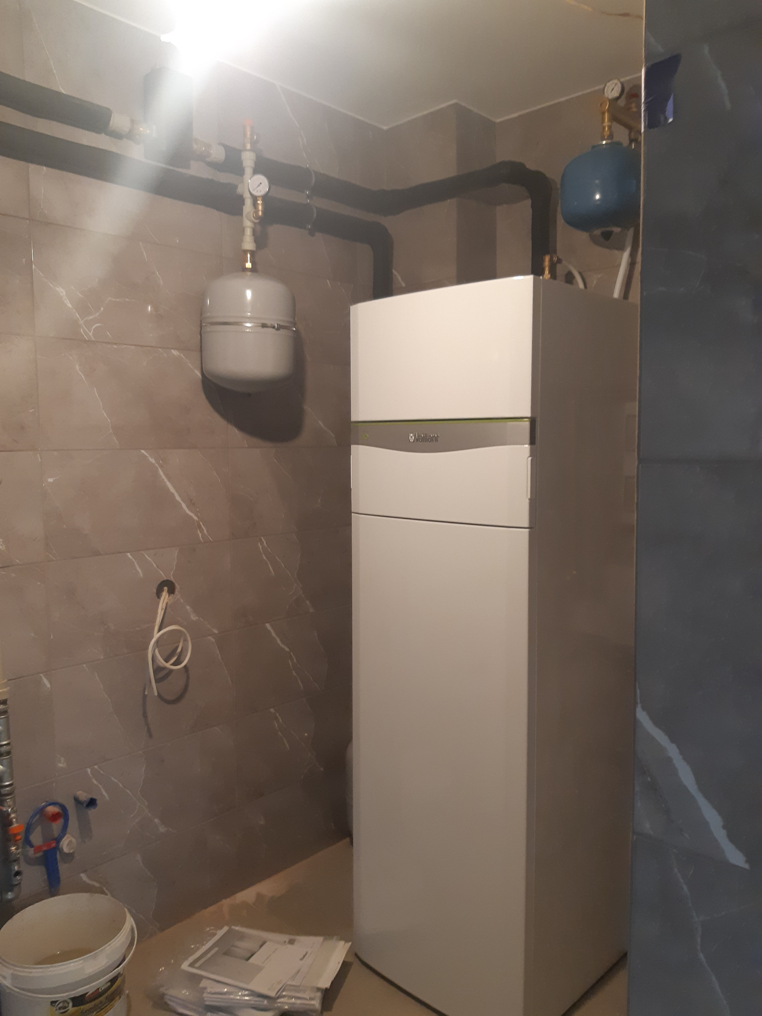 Nowoczesny, biały piec gazowy Vaillant zainstalowany w pomieszczeniu z szarymi płytkami imitującymi marmur, widoczne elementy instalacji CO, rury, zbiornik wyrównawczy i manometry, w tle ściana...