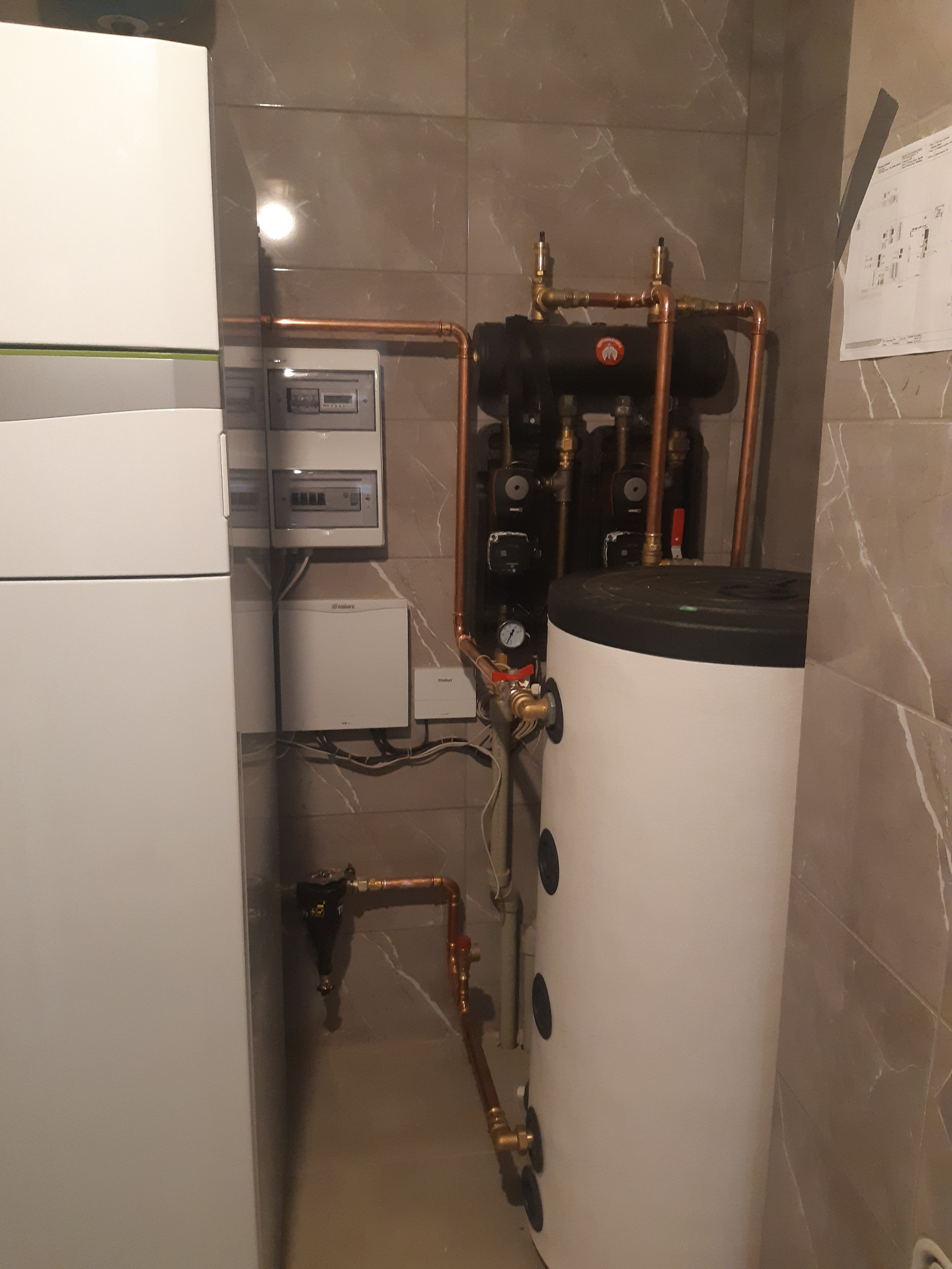 Nowoczesna kotłownia z piecem gazowym, zasobnikiem ciepłej wody, rozdzielaczem hydraulicznym i instalacją z miedzianych rur na tle płytek imitujących marmur.