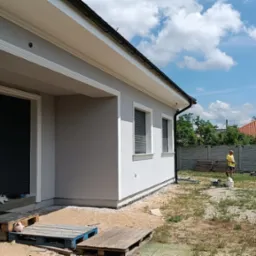 Nowoczesny dom jednorodzinny z szarą elewacją i białymi zdobieniami okiennymi oraz podbitką dachową, widoczny pracownik w oddali, teren wokół w trakcie prac wykończeniowych.