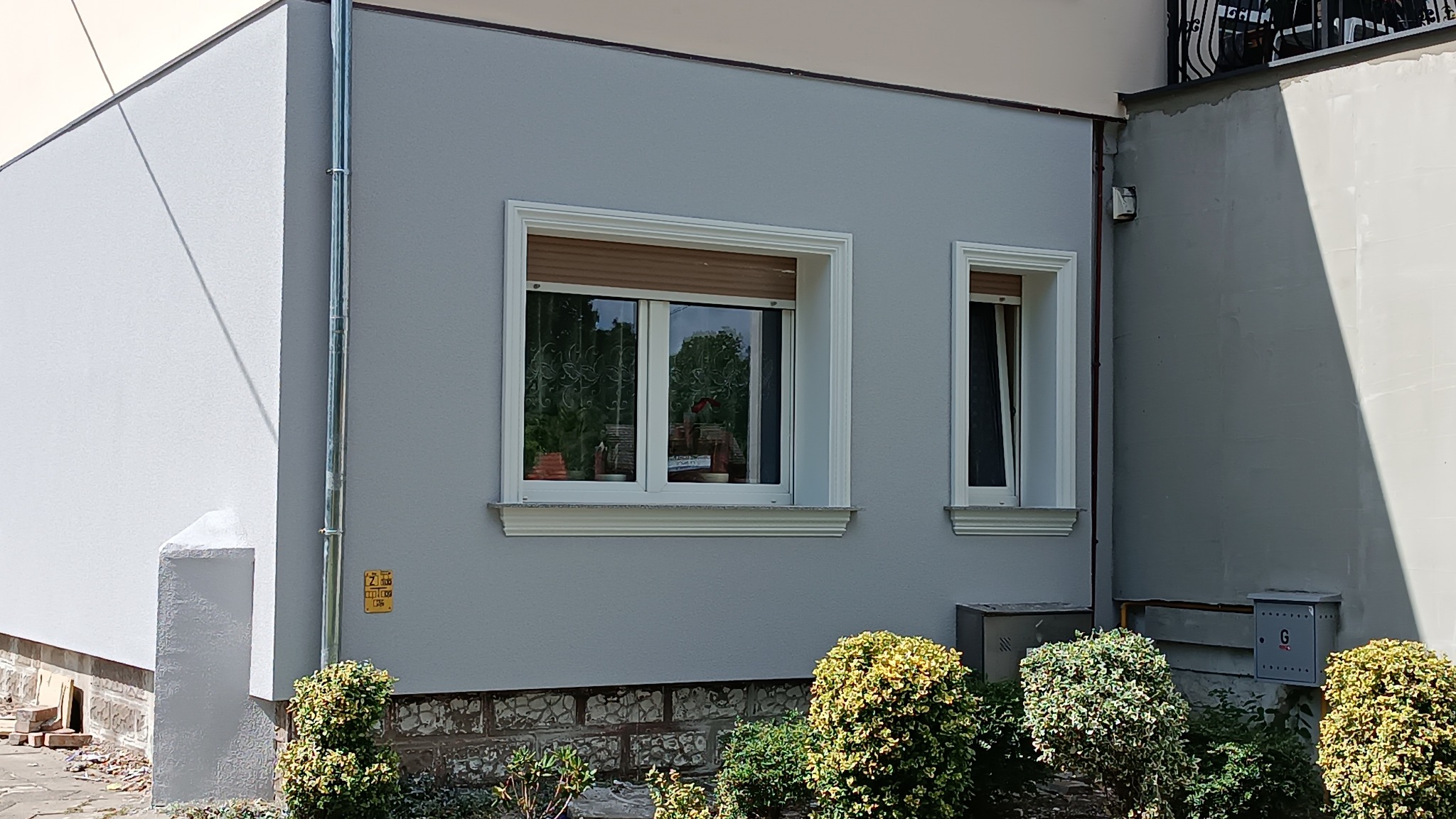 Jasnoszara elewacja budynku po remoncie w Ośnie Lubuskim, widoczne dwa okna z białymi ramami i ozdobnymi gzymsami, krzewy u podstawy ściany.