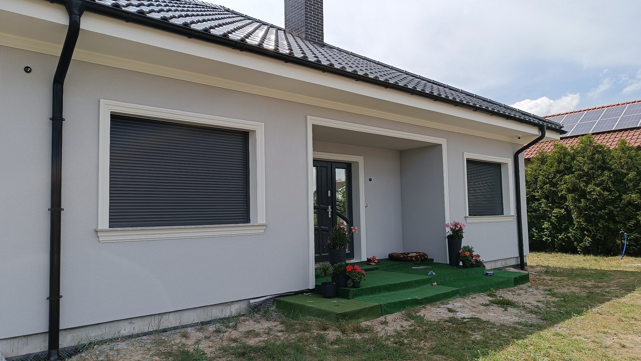 Jasnoszary dom jednorodzinny z czarną dachówką i roletami zewnętrznymi, widok z zewnątrz na elewację z dekoracyjnymi listwami i zielonymi schodami wejściowymi.