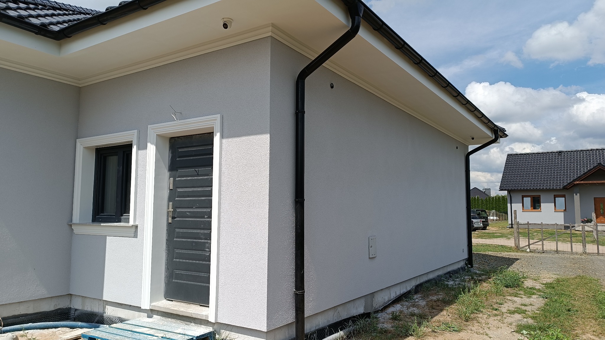 Nowoczesny dom jednorodzinny z szarą elewacją o strukturze baranka, czarną stolarką okienną i drzwiową z białymi obramowaniami, oraz czarnymi rynnami w słoneczny dzień.