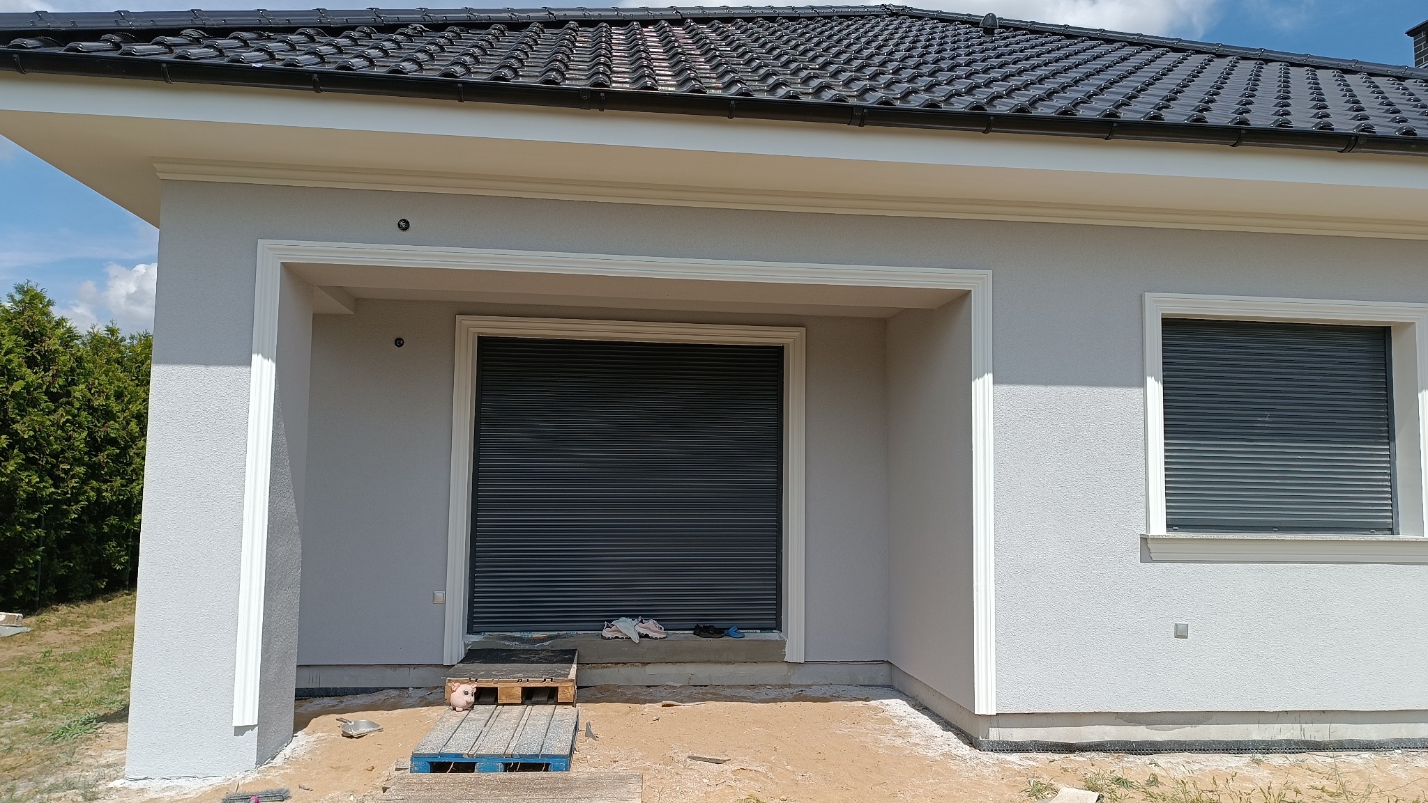 Jasnoszara elewacja domu jednorodzinnego z czarną dachówką, ozdobnymi białymi listwami wokół okien i wnęki garażowej z zamkniętą roletą, widoczne ślady prac budowlanych na podłożu.