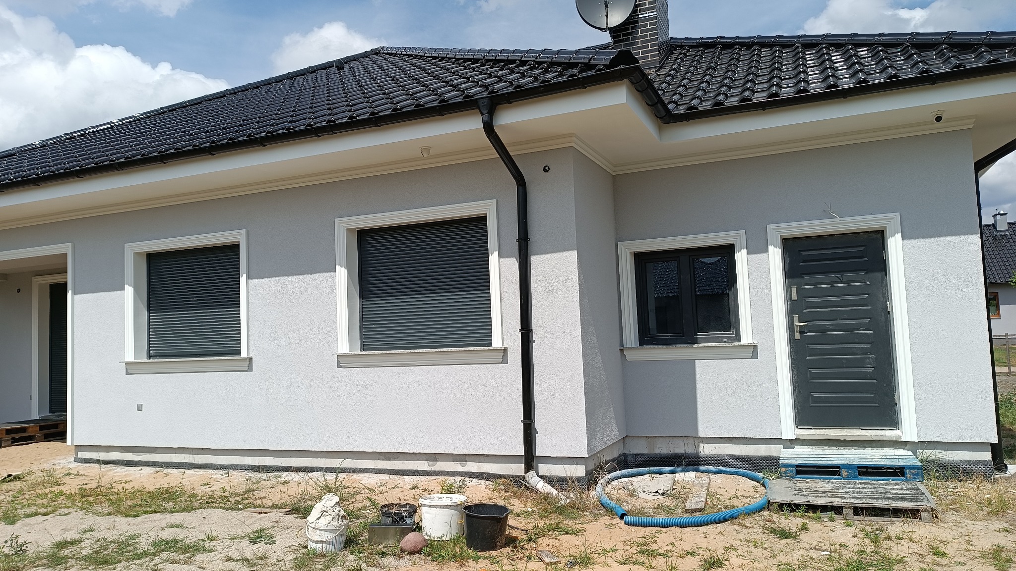 Szary dom jednorodzinny z czarnym dachem i oknami z roletami zewnętrznymi, widoczny fragment elewacji z detalami sztukaterii okiennej i drzwiowej, rynna oraz teren wokół budynku w trakcie prac...