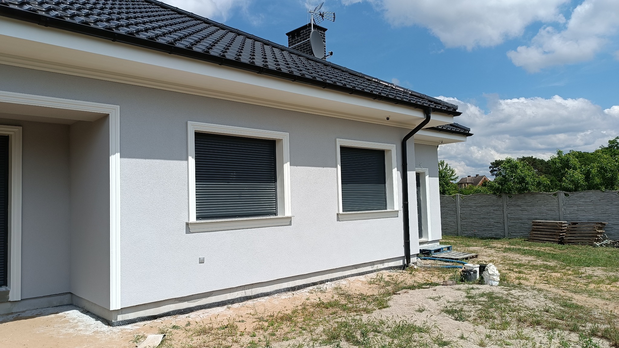 Jasnoszary dom jednorodzinny z czarną dachówką i roletami zewnętrznymi, widoczne detale wykończeniowe elewacji i przygotowane materiały budowlane na trawniku.