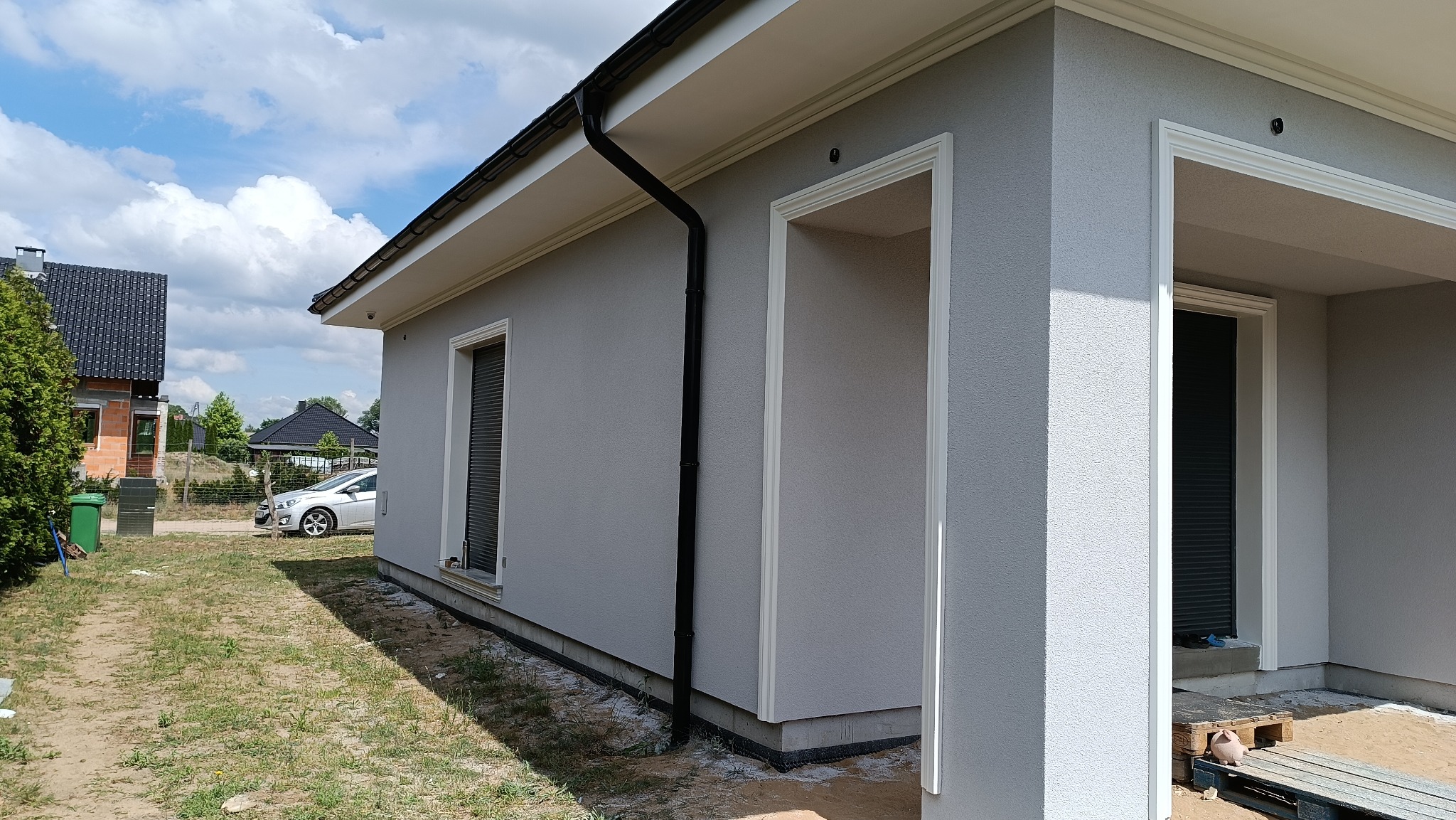 Jasnoszara elewacja domu jednorodzinnego z białymi obramowaniami okien i drzwi, widoczny fragment dachu z rynną, w tle inne domy i błękitne niebo z chmurami.