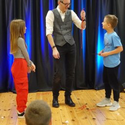Iluzjonista Piotr Fijka - Mężczyzna w okularach i kamizelce prezentuje trik z czerwoną liną dwójce dzieci na scenie z ciemnym tłem i niebieskim oświetleniem.