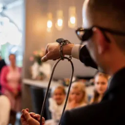 Iluzjonista prezentuje trik z łańcuchem i metalową obręczą na tle publiczności, w tle widoczne lampy i kwiaty, ujęcie zza pleców artysty.