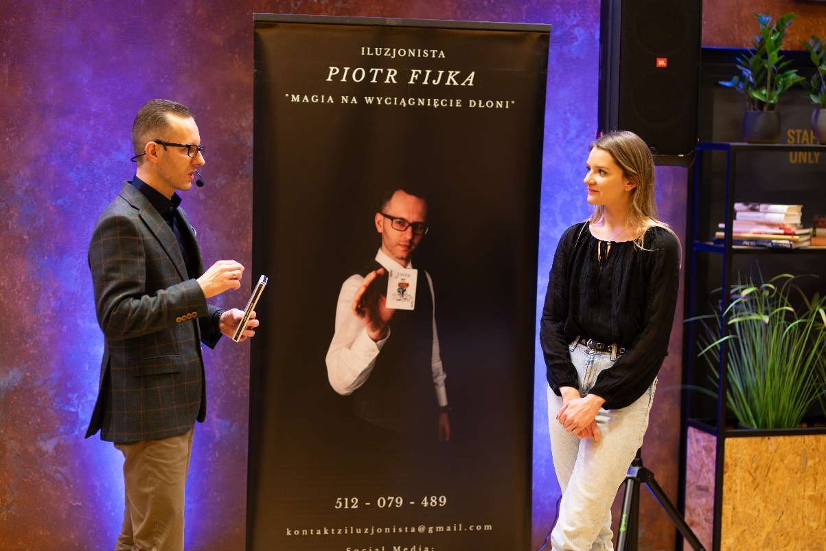 Iluzjonista w trakcie występu z mikrofonem, obok baner reklamowy z wizerunkiem iluzjonisty prezentującego kartę Joker oraz kobieta obserwująca pokaz.