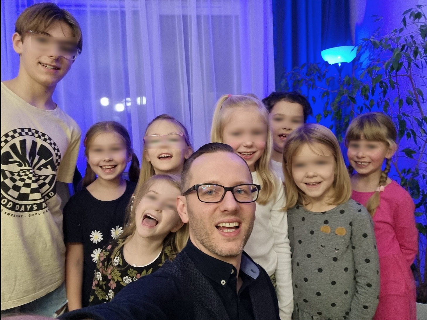 Zdjęcie grupowe: uśmiechnięty mężczyzna w okularach robi selfie z grupą roześmianych dzieci na tle niebieskich zasłon i rośliny doniczkowej.