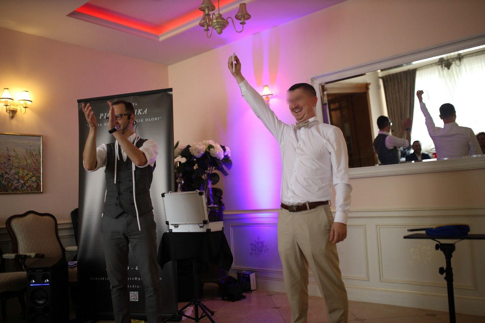 Dwóch mężczyzn w trakcie występu iluzjonistycznego; jeden w kamizelce i okularach klaszcze, drugi w koszuli i muszce trzyma coś w uniesionej dłoni, w tle białe kwiaty i lustro odbijające scenę.