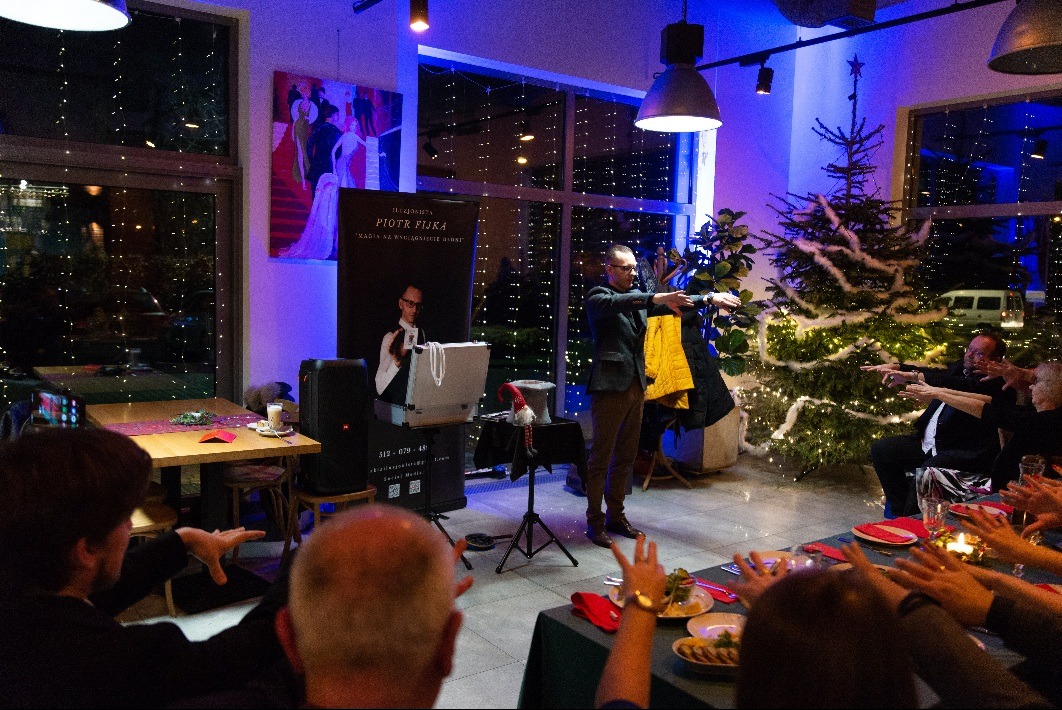 Mężczyzna w marynarce wykonuje gest iluzjonistyczny przed grupą osób siedzących przy stole w pomieszczeniu z dekoracjami świątecznymi i plakatem reklamowym z wizerunkiem iluzjonisty w tle.