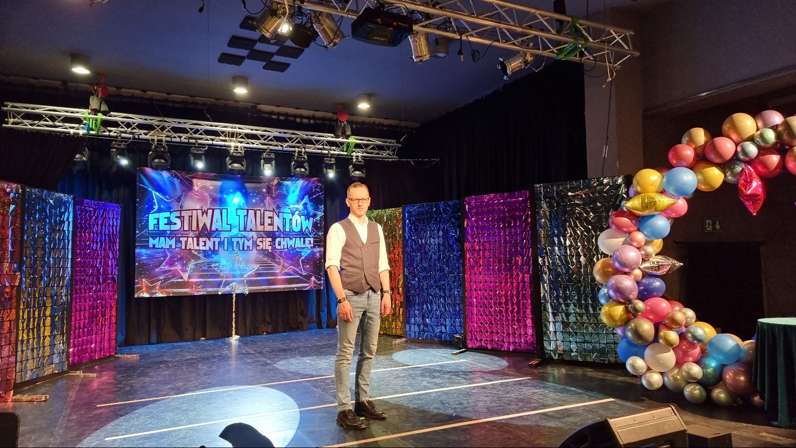 Iluzjonista w okularach i kamizelce na scenie przed ekranem 'Festiwal Talentów' i dekoracjami z balonów.