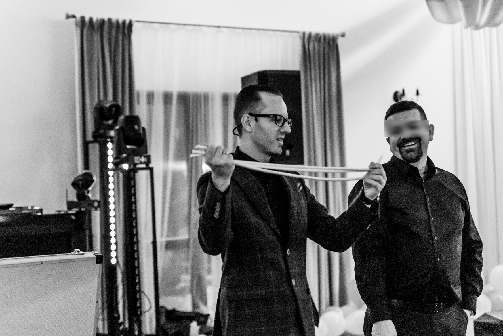 Iluzjonista w kraciastej marynarce prezentuje trik z połączonymi białymi sznurkami, uśmiechnięty widz z zamazaną twarzą obserwuje występ, w tle oświetlenie sceniczne i białe balony.