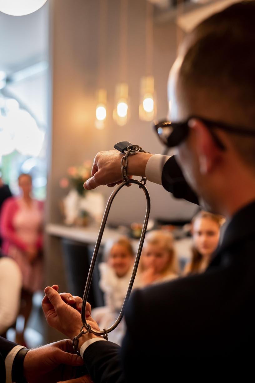 Iluzjonista prezentuje trik z łańcuchem i metalową obręczą na tle publiczności, w tle widoczne lampy i kwiaty, ujęcie zza pleców artysty.