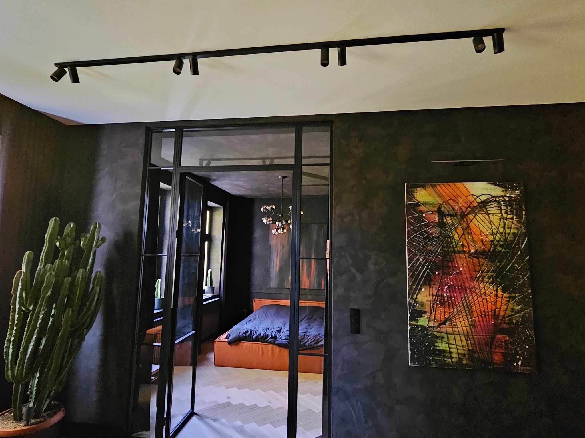 Elegancka sypialnia za szklaną ścianą z czarnymi profilami. Ciemne ściany z fakturą, abstrakcyjny obraz, oświetlenie szynowe i duży kaktus. Parkiet w jodełkę.