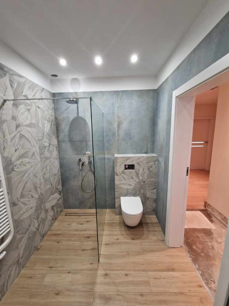 Nowoczesna łazienka z kabiną typu walk-in, płytkami w odcieniach szarości i drewna, oraz podtynkową toaletą. Widoczne oświetlenie punktowe i minimalistyczny design.