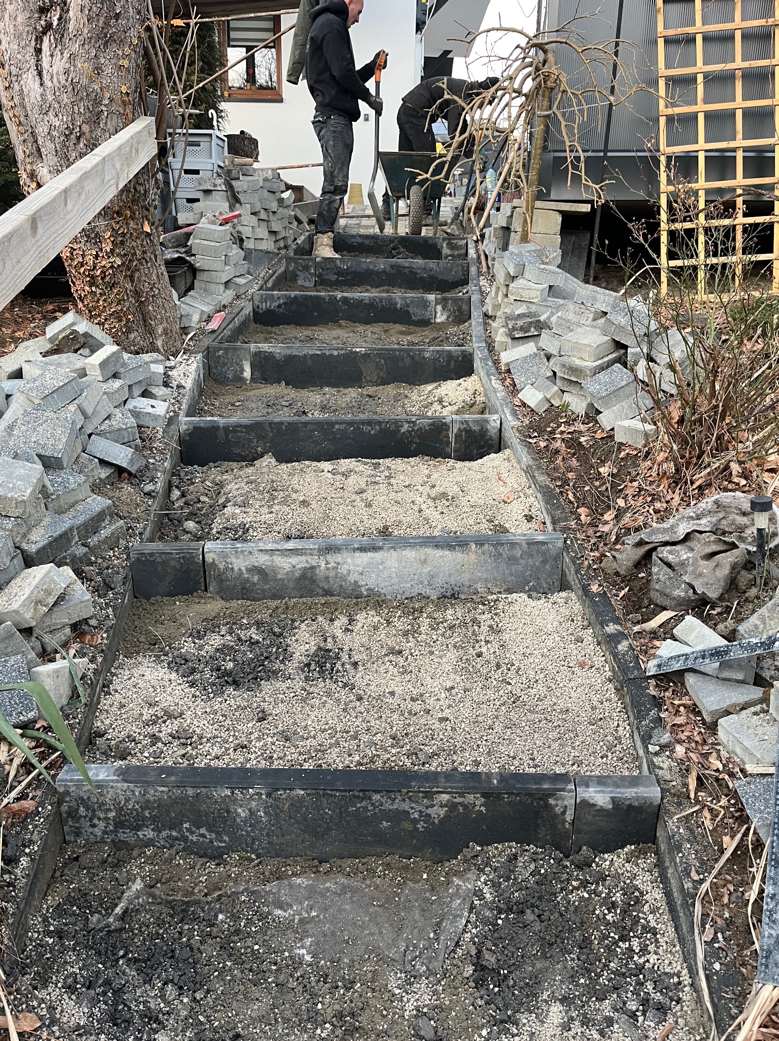 Budowa schodów zewnętrznych z czarną obwódką i wypełnieniem z szarego kruszywa, pracownicy w tle, stosy kostki brukowej po bokach, widok z góry na plac budowy w ogrodzie.