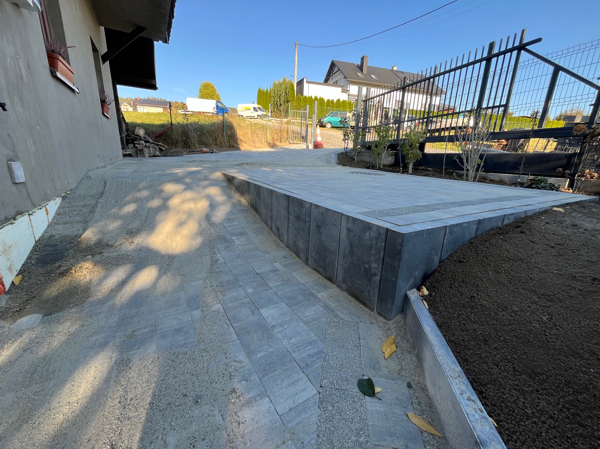 Ukończony podjazd z szarej kostki brukowej, obrzeża z ciemnych betonowych bloków, świeżo nawieziona ziemia ogrodowa, widok na dom i ulicę w słoneczny dzień.