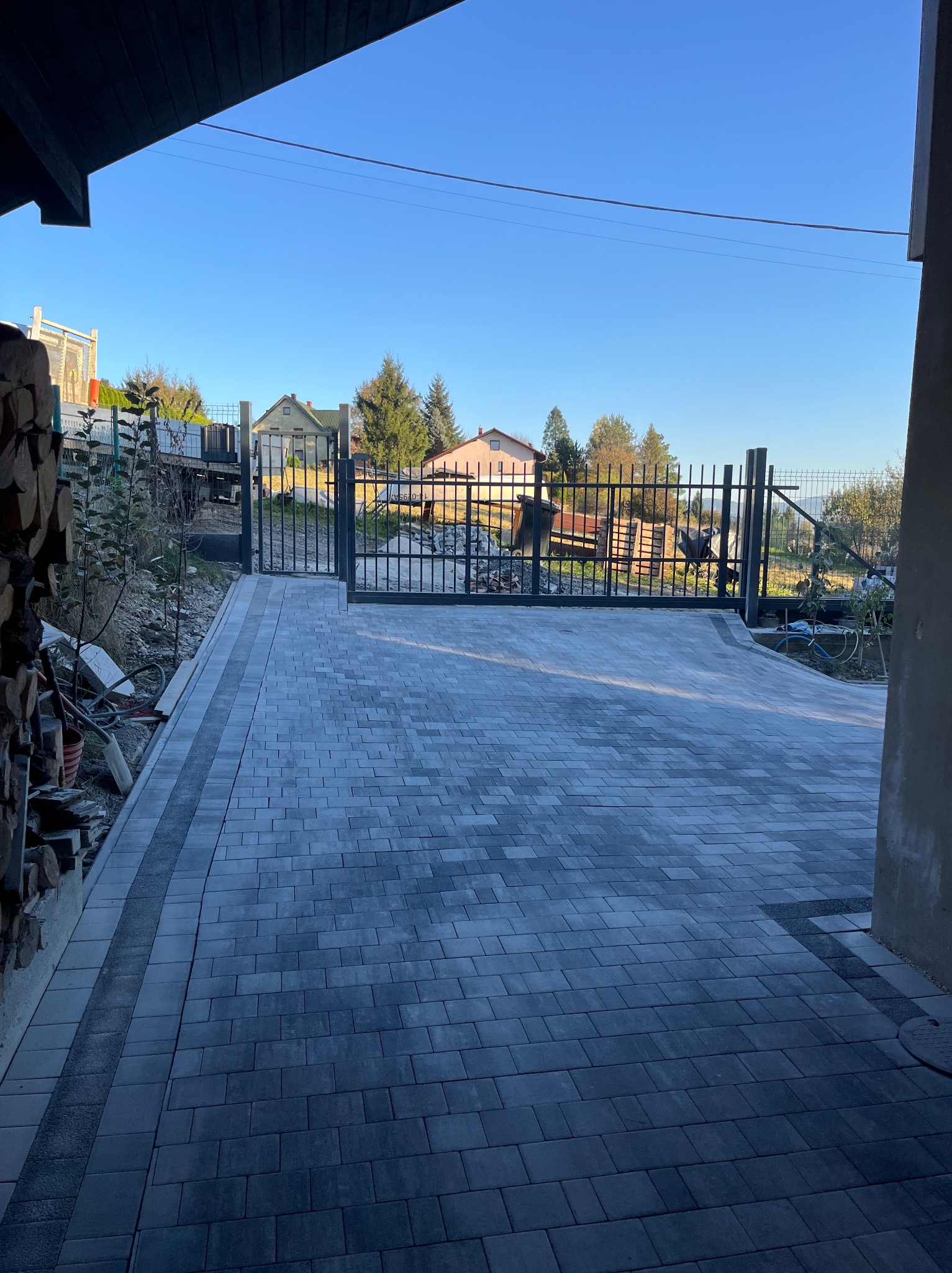 Ułożona szara kostka brukowa prowadząca do czarnej bramy wjazdowej, widok z zadaszonego tarasu, w tle domy i błękitne niebo.